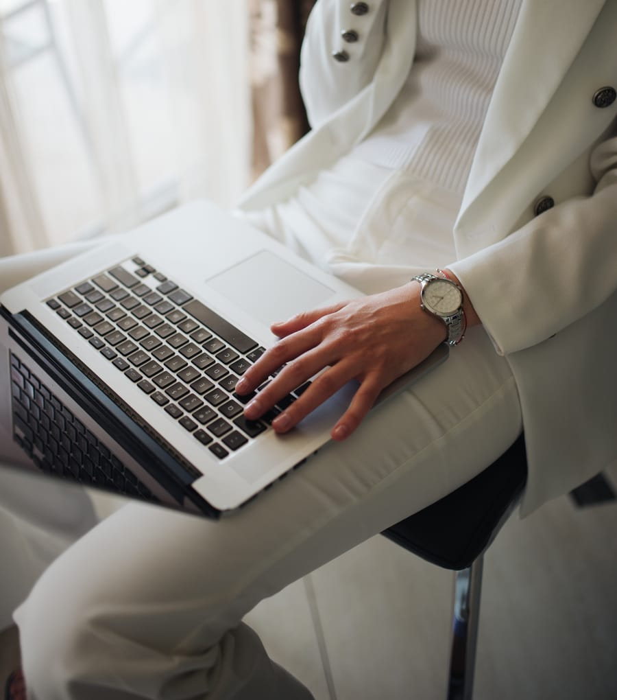 A person in a white suit working on a laptop balanced on one knee beside a bright window, face out of frame.