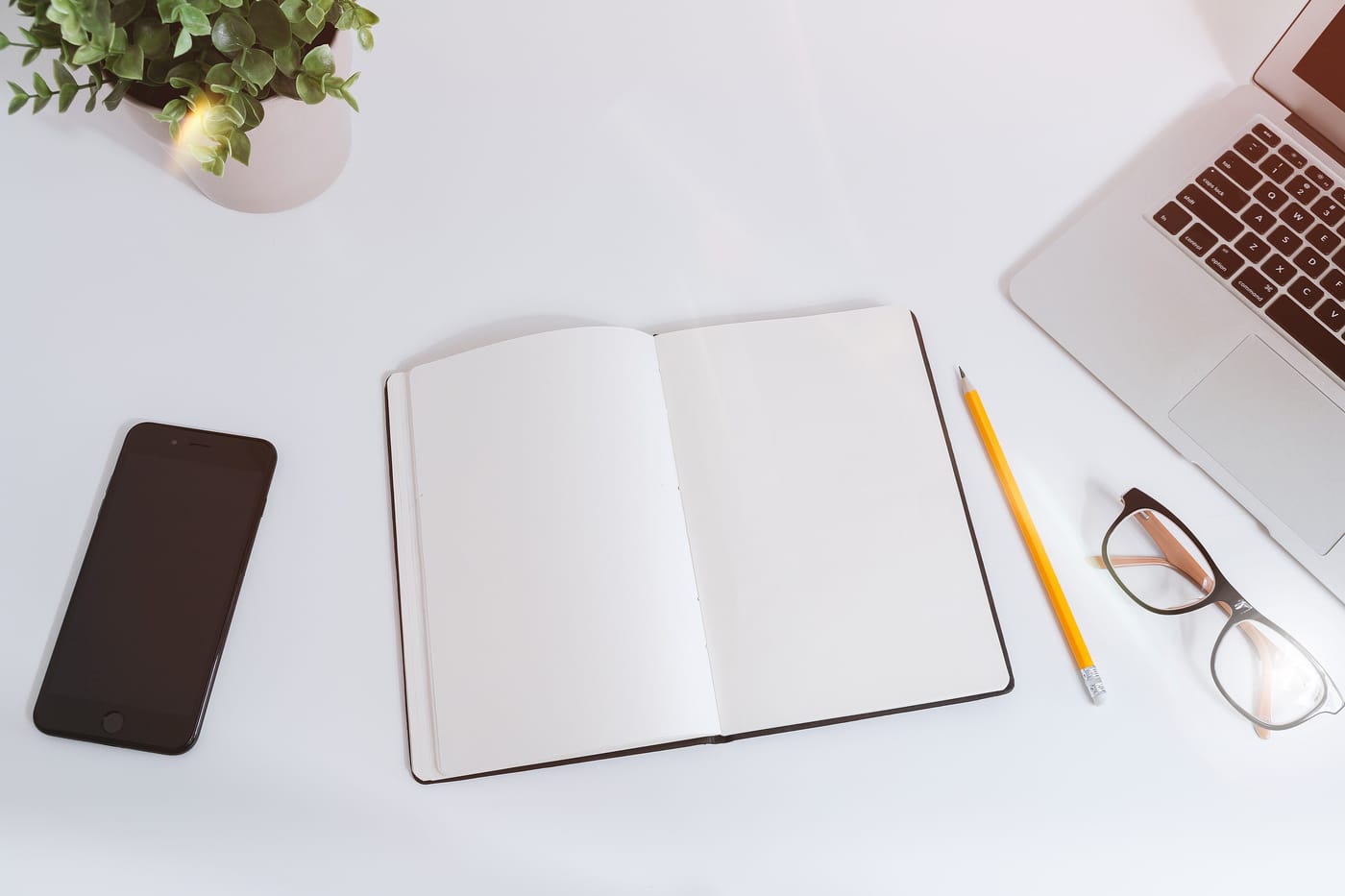 An open blank notebook, a yellow pencil, a pair of glasses and a black phone on a white desk beside a laptop.