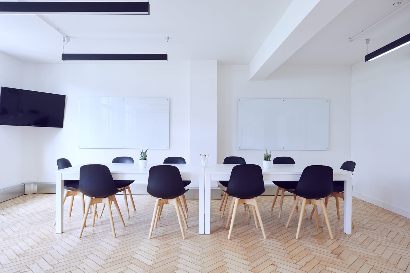 A bright meeting room with white tables, navy chairs on wooden legs and two whiteboards on a white wall.