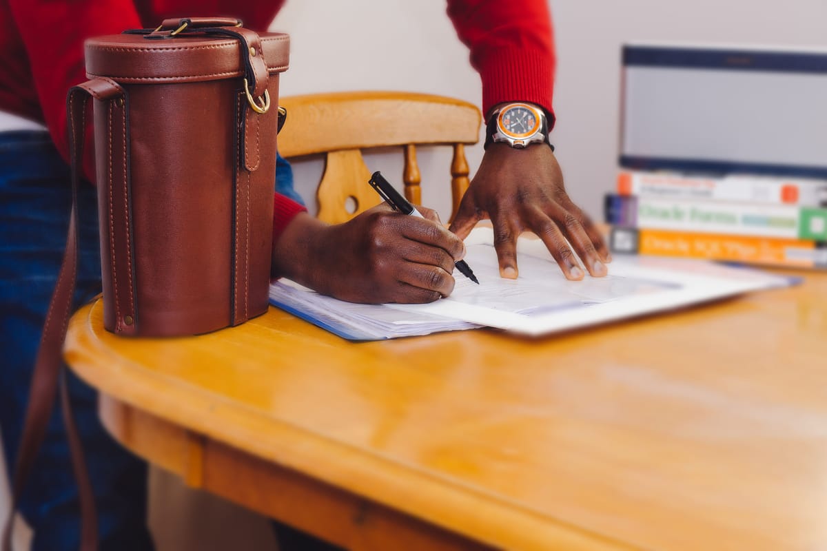 A man writing notes on a printed page at a wooden desk, a brown leather case beside his hand.