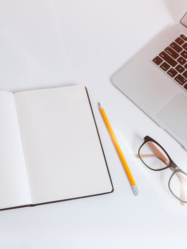 A desk laid out with an open notebook, pencil, reading glasses and a laptop