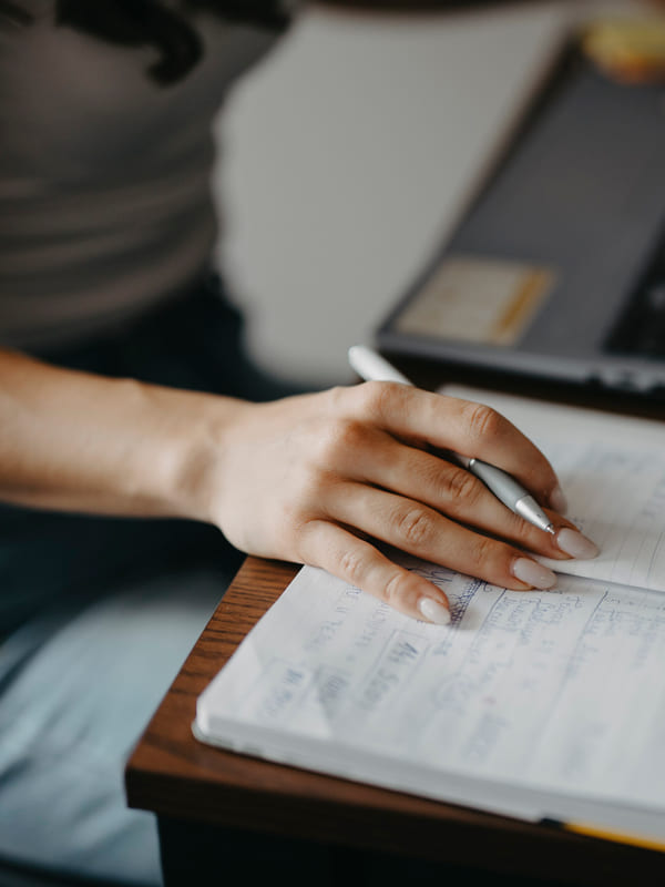 Notes being written in a notebook beside an open laptop on a desk