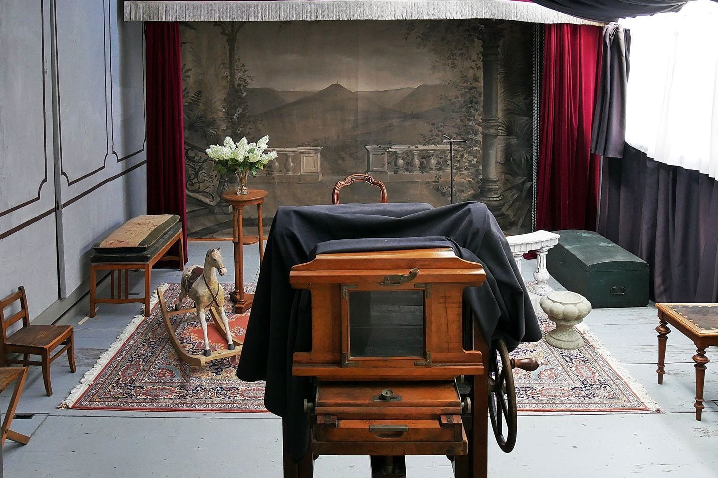 A painted scenic backdrop and a wooden plate camera under its focusing cloth in a historic portrait studio.