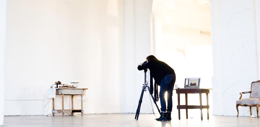 A sitting under way in a white room: the photographer bent to the camera on its tripod, a tethered laptop open on the table beside him and the chair waiting in the light