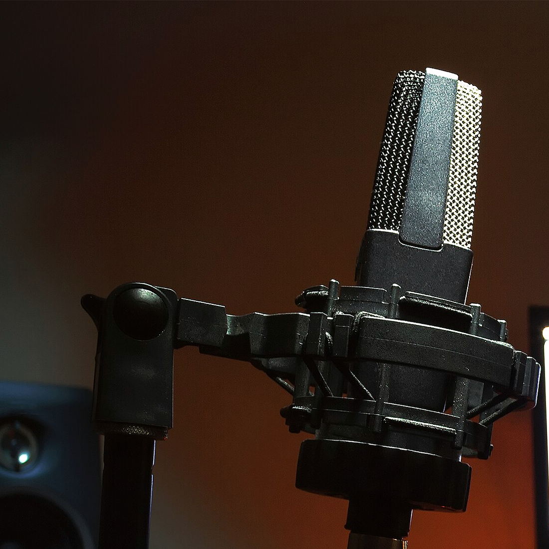 A condenser microphone in a shock mount on a stand against a warm brown wall, beside the edge of a blue-lit monitor.