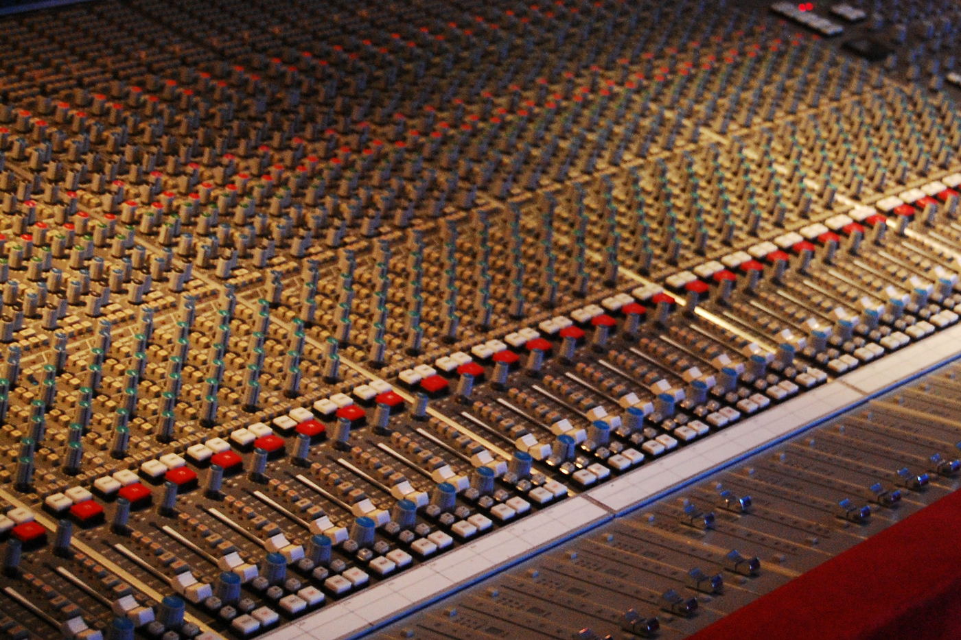 A very large bank of faders in receding perspective, with red-trimmed channel strips above a red leather armrest.