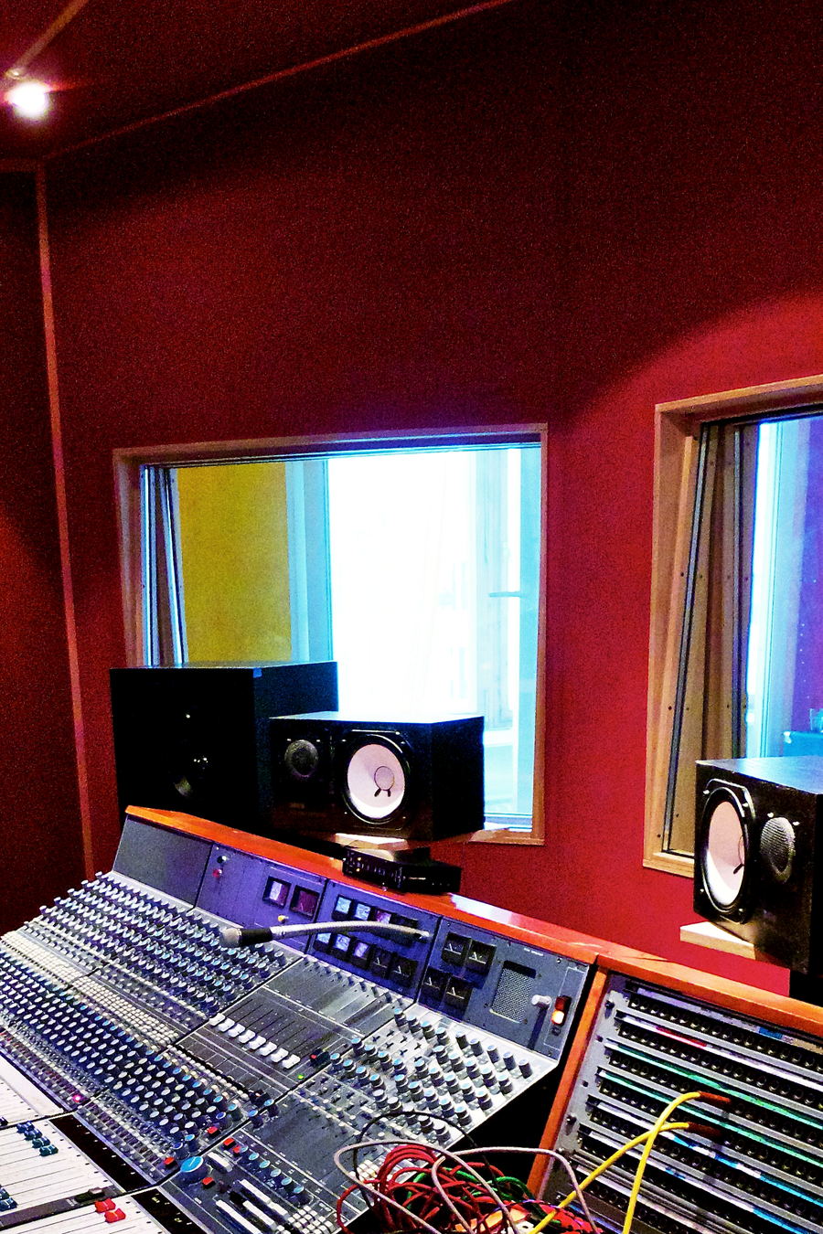 A vintage mixing console in a red-walled control room, with monitors on the bridge, patch cables and two windows behind.