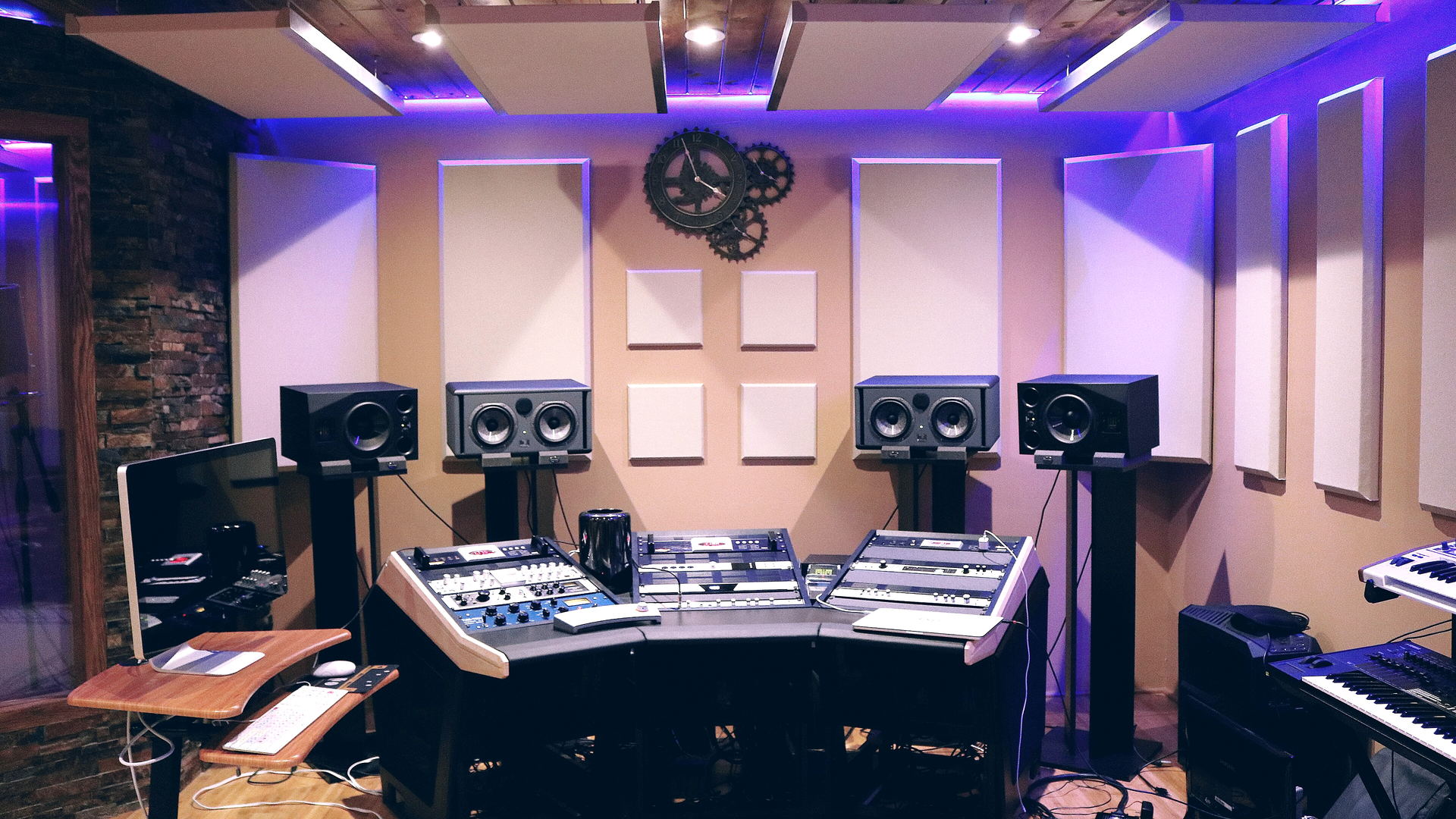 A control room under a purple-lit ceiling, with cream acoustic panels, a gear-shaped wall clock and four monitors over an angled desk of outboard units.