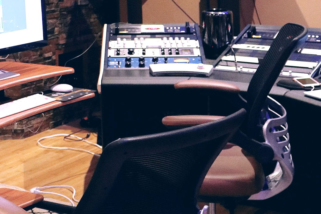 An angled desk of blue outboard units beside a black mesh chair with a wooden armrest, above a wooden floor.