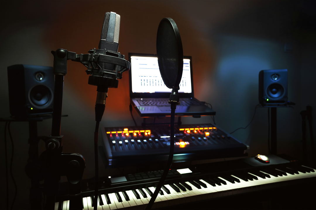 A large-diaphragm condenser microphone in a shock mount with a pop filter, in front of a dark desk with a laptop and a lit controller.