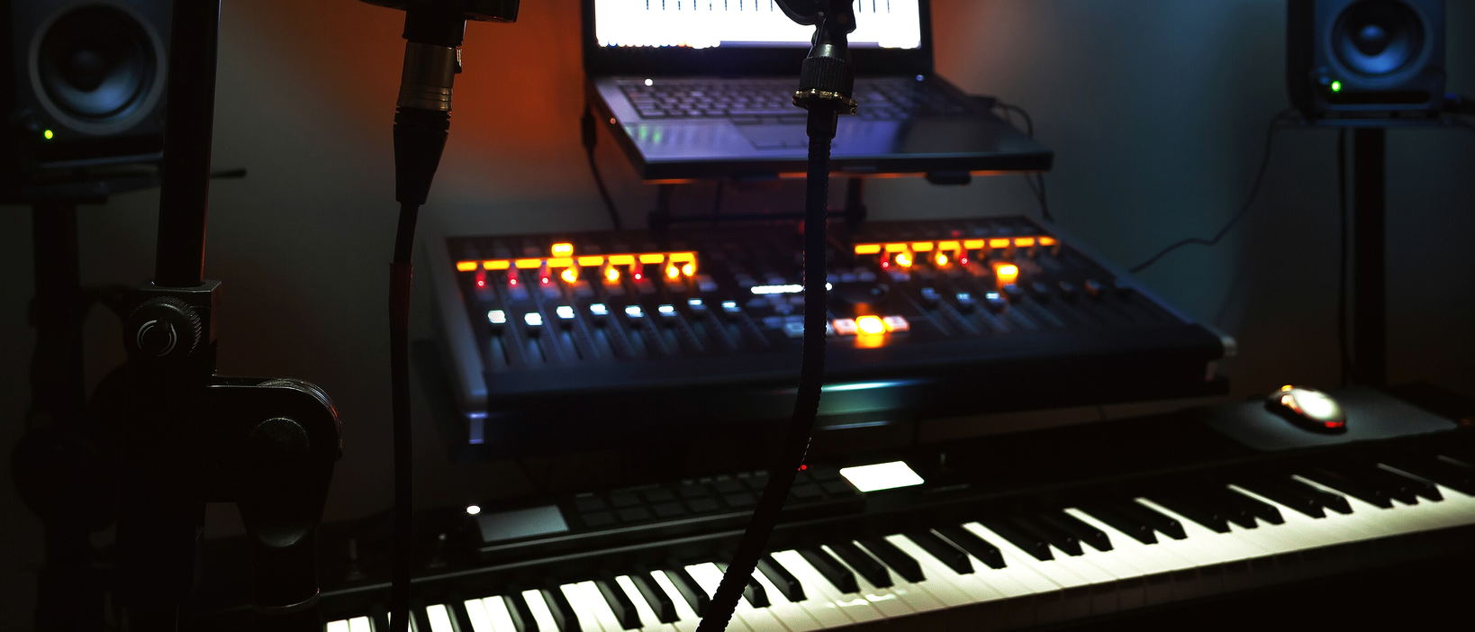 A microphone stand in the foreground, a lit controller, a keyboard, a laptop and monitors in a dark room