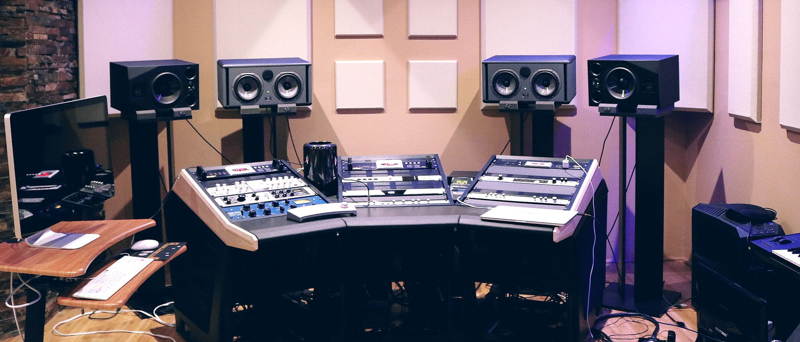 A control room under purple light, with four monitors on stands and an angled desk of outboard units between cream acoustic panels.