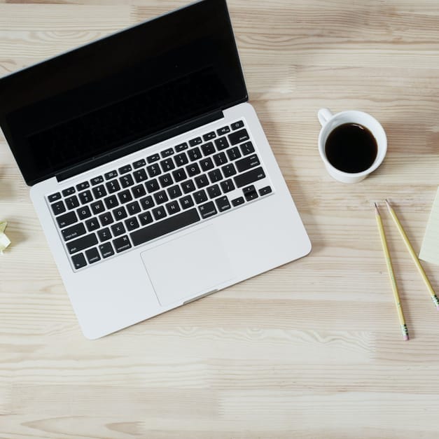 A laptop, a cup of coffee, a notepad and two pencils arranged on a pale wooden desk.