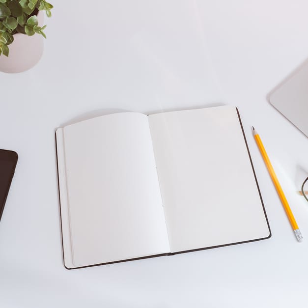 An open notebook, a pencil, reading glasses and a phone arranged beside a laptop on a white desk.