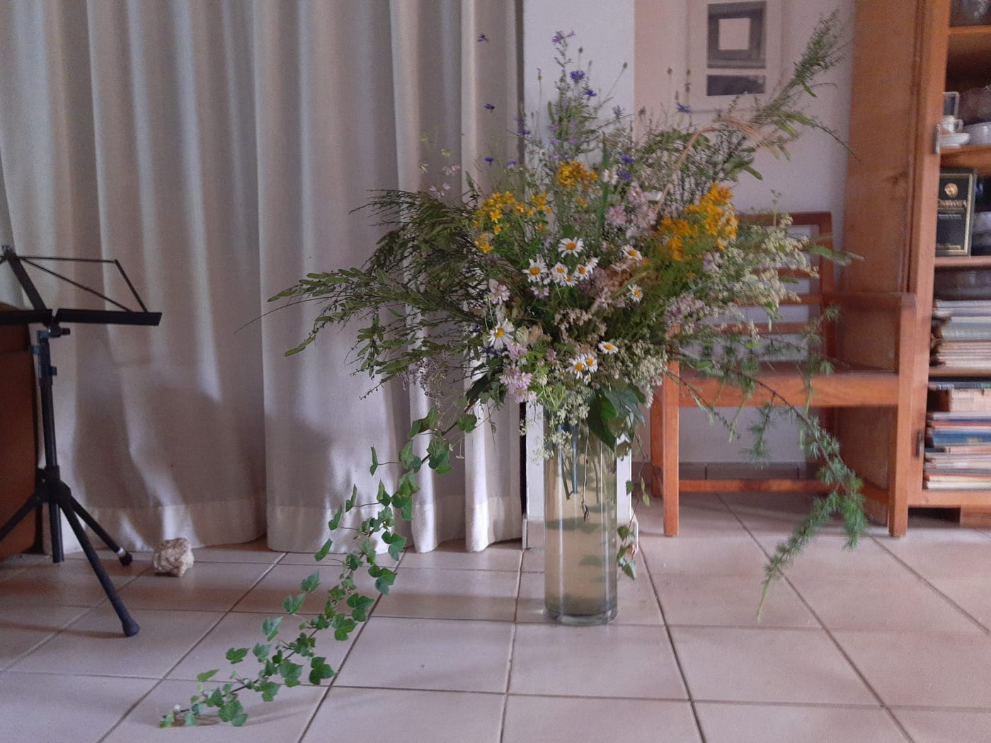 Wildflowers arranged in a tall clear glass vase standing on a tiled floor.