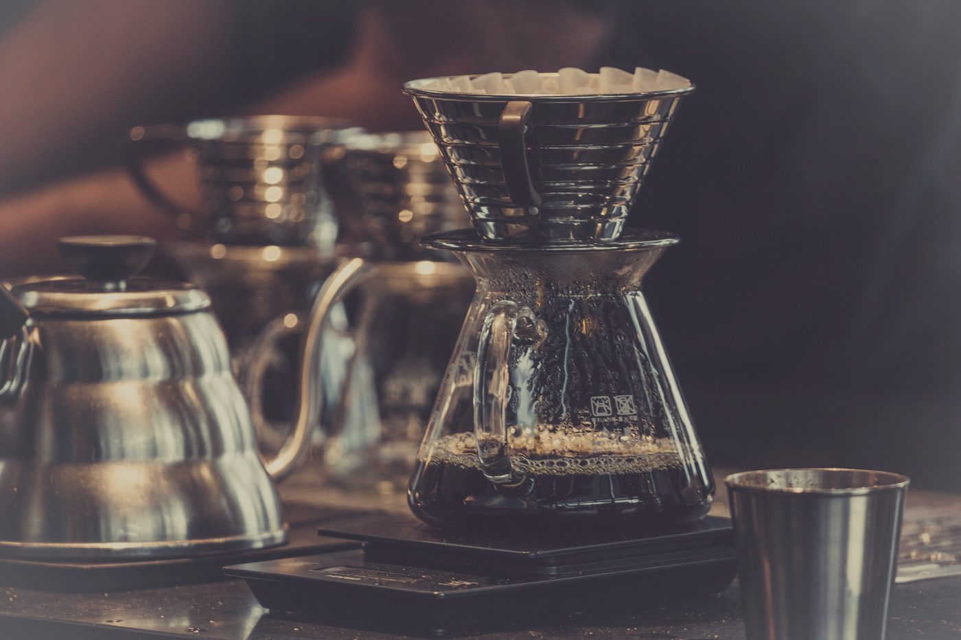 A pour-over cone and glass carafe standing on a counter with two gooseneck kettles and a small metal cup beside them.