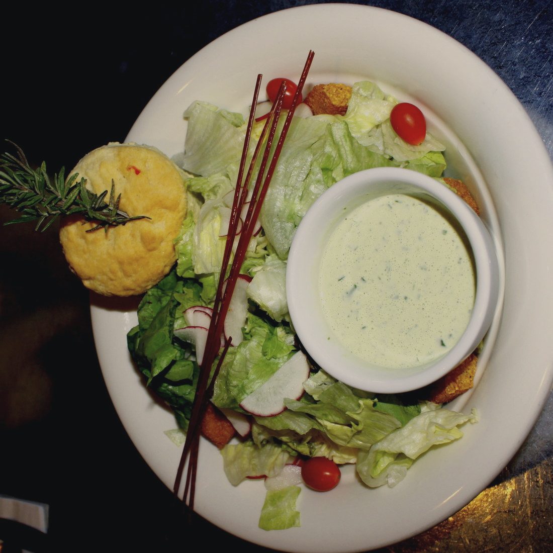 A plate of salad leaves, radish and tomato around a small pot of pale green dressing, with a soft roll at the edge.