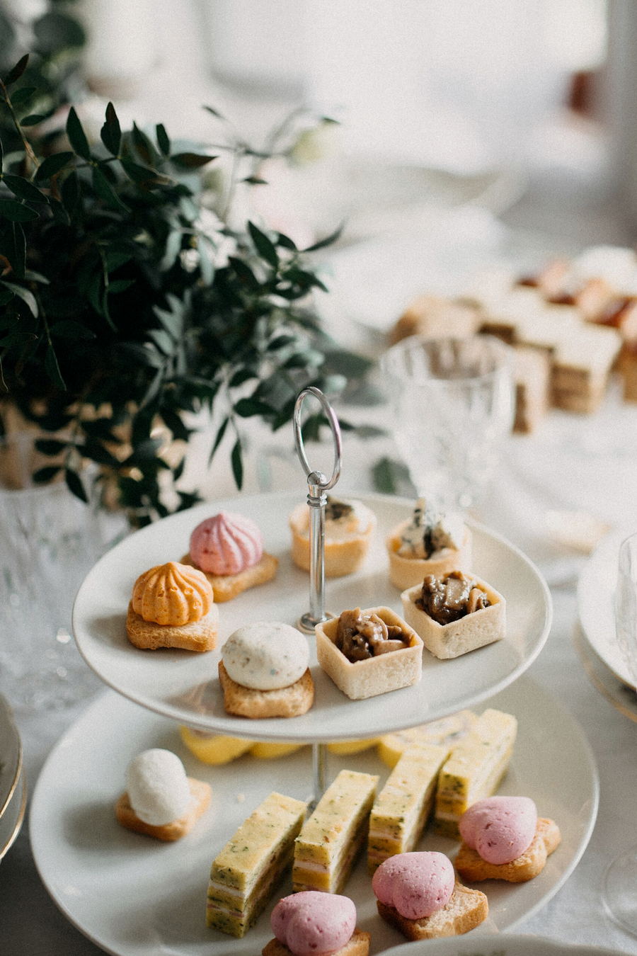 A two-tier china stand of meringues, tartlets and small sandwiches beside cut-crystal glasses and green foliage on a white cloth.