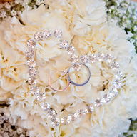 A diamante heart and two rings lying on a bouquet of cream carnations.