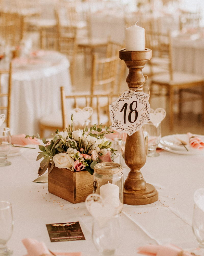 A wooden box of cream roses and eucalyptus beside a turned candlestick carrying table number 18, on a white cloth laid with pink napkins.
