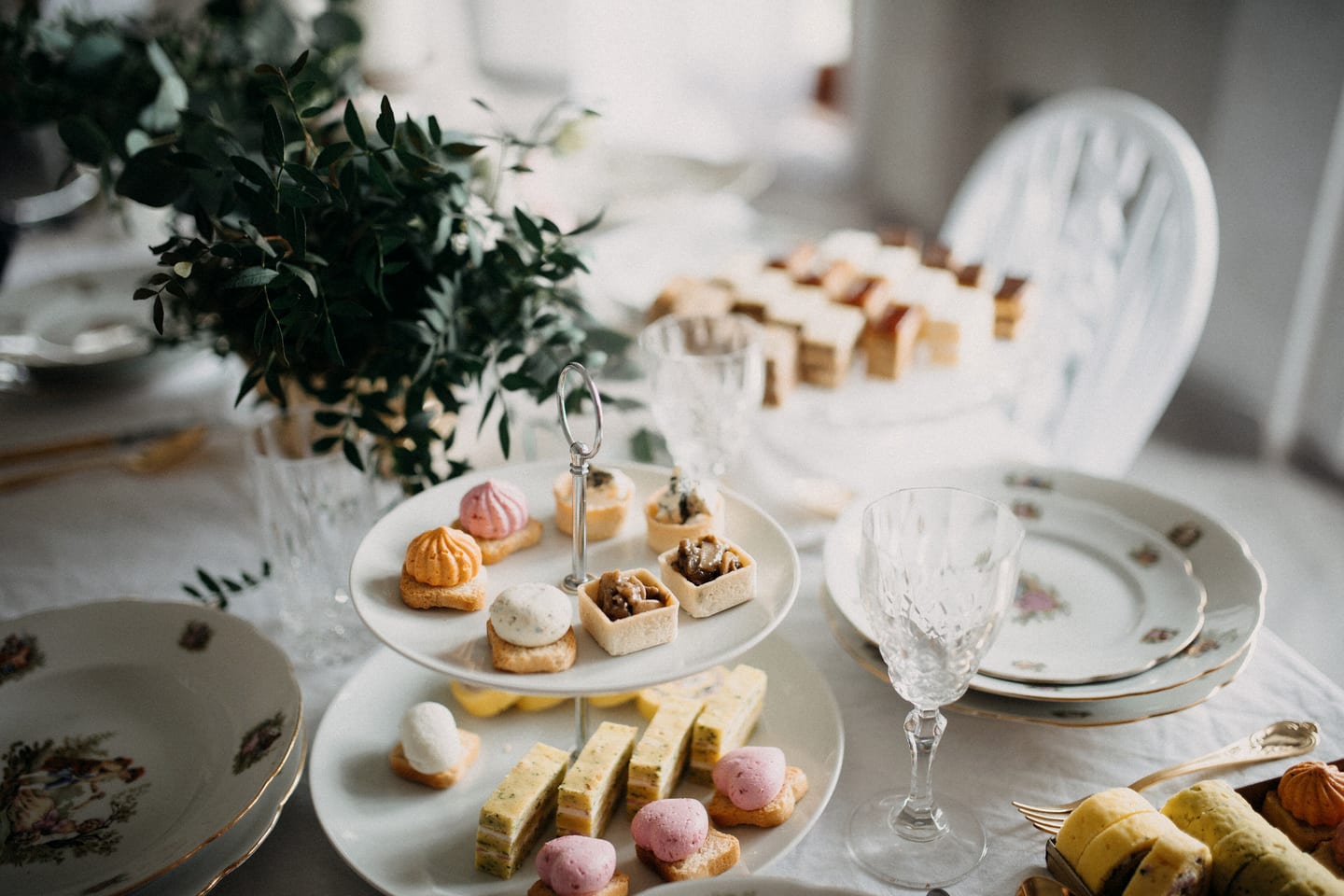 A two-tier china stand of meringues, tartlets and small cakes beside a cut-crystal glass on a white cloth.