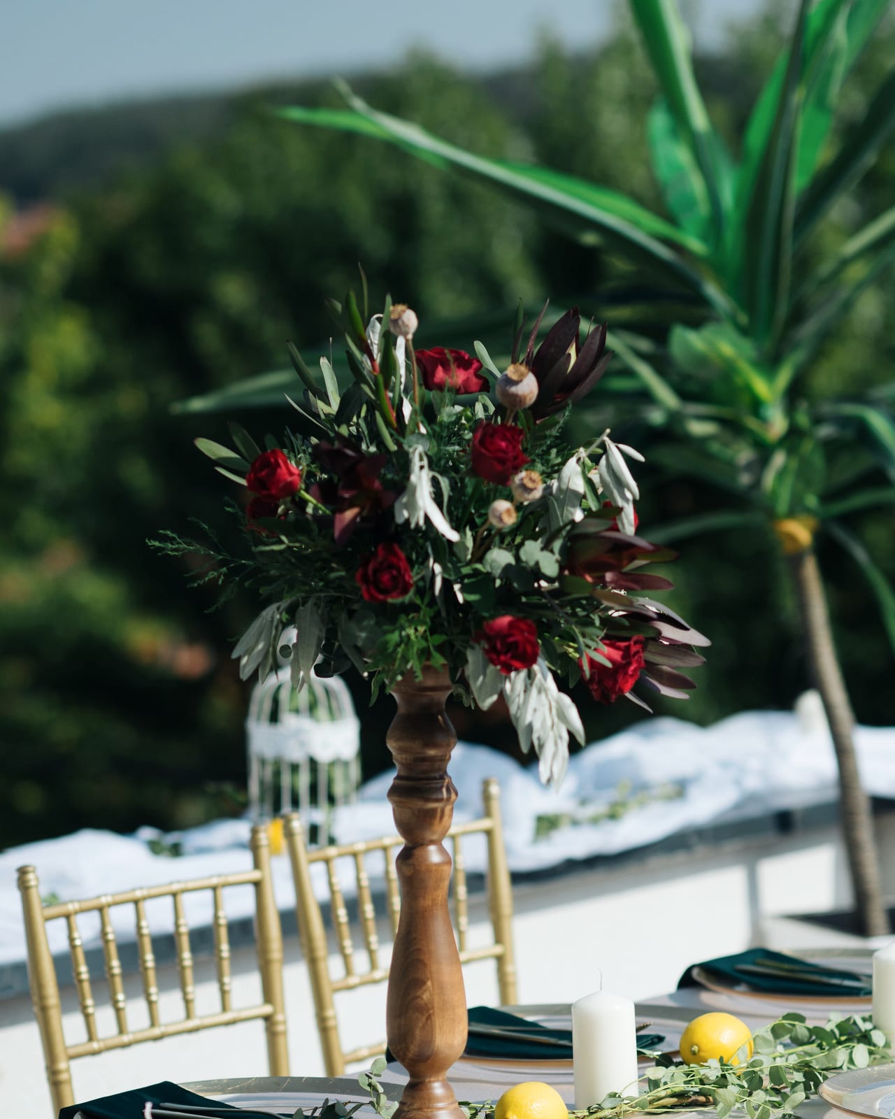 A tall arrangement of dark red roses and grey-green foliage on a turned wooden stand, set outdoors above gold chairs.