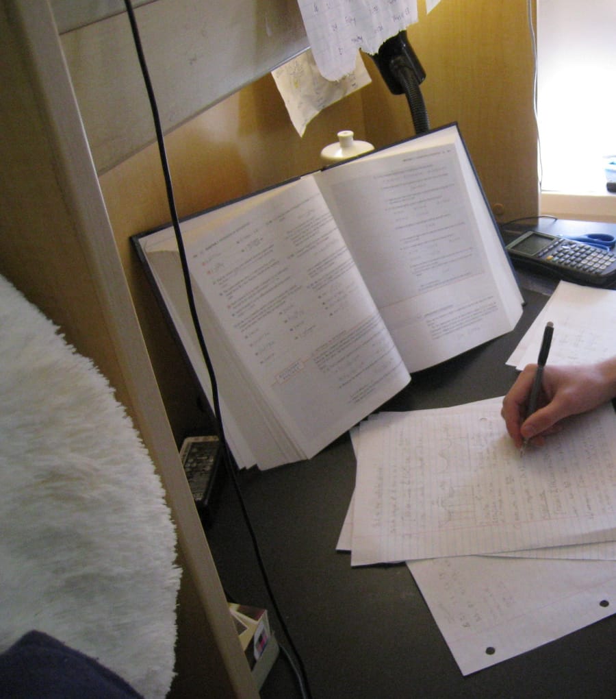 A hand writing notes on lined paper beside an open textbook and a calculator on a small study desk.