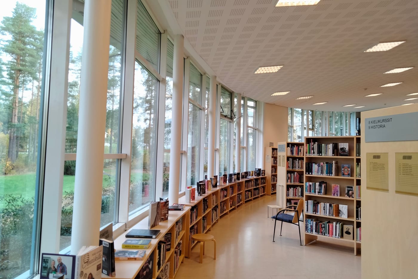 A long library room with low bookshelves running beside floor-to-ceiling windows that look out on pine trees.