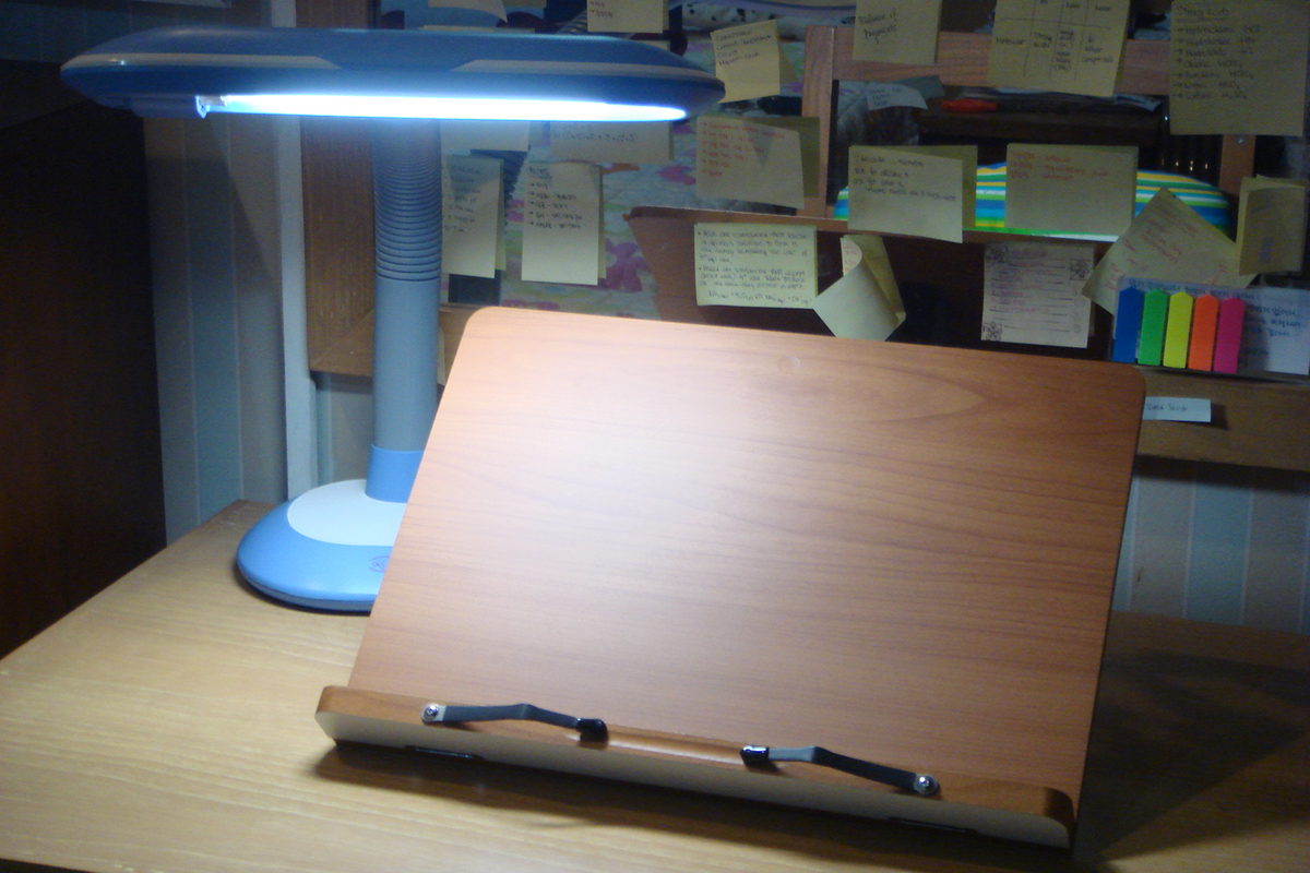 A wooden bookstand and a lit desk lamp on a study desk, with a board of handwritten revision notes on cards behind them.