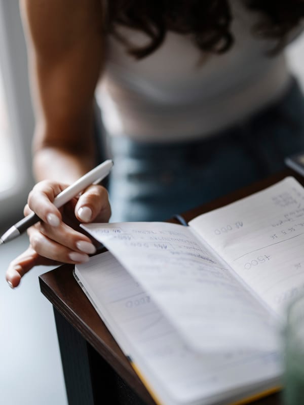 Hands writing longhand notes in an open exercise book by a window